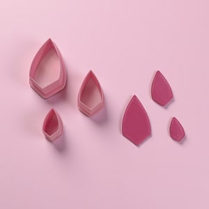 May include: A collection of pink petal-shaped cookie cutters and cut-out petals on a pink background. The cutters are in various sizes, with a layered design. The cut-out petals are solid pink and also vary in size.