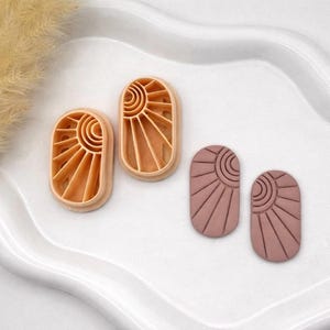 May include: Two sets of sun-shaped clay cutters and two sun-shaped clay pieces. The cutters are light brown, and the clay pieces are a dusty rose color. The sun design features a circular center with radiating lines.