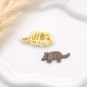 May include: Two armadillo-shaped items. One is a light yellow and white cookie cutter with a detailed armadillo design. The other is a small, gray armadillo figurine with striped detailing on its back.