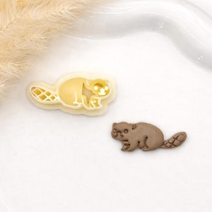 May include: Beaver-shaped cookie cutter and a brown beaver-shaped fondant decoration. The cookie cutter is yellow and white, and the fondant beaver is brown. The items are on a white surface.