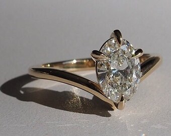 Può includere: Un anello d'oro con un grande diamante di forma ovale incastonato a griffe. La fascia è sottile e si curva verso l'alto per incontrare il diamante.
