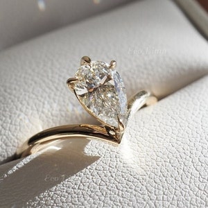 May include: A pear-shaped diamond engagement ring set in a gold band with a V-shaped design. The ring is displayed in a white ring box.