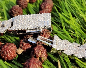 Op de afbeelding: Een close-up van een zilveren ketting met een diamant-ingelegd ontwerp. De ketting ligt op een bed van bruine kralen en groen gras.