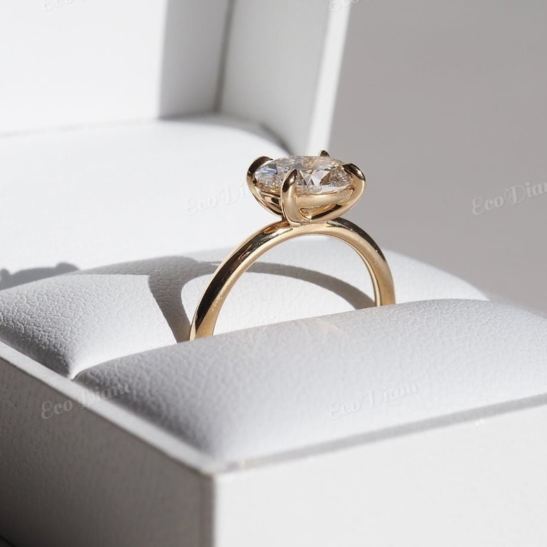 May include: A gold engagement ring with a large, clear oval-shaped diamond set in a simple, elegant band. The ring is displayed in a white ring box.