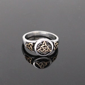 Puede incluir: Un anillo de plata con un diseño de nudo celta dorado. El anillo es una banda simple con un diseño circular en relieve en el centro.