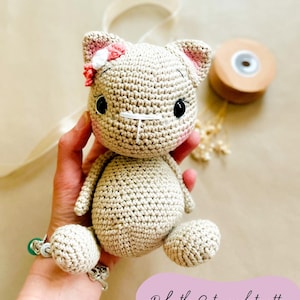 May include: A crocheted cat doll in beige yarn, with pink accents on the ears and a small flower. The cat has black eyes and a stitched nose and mouth. The text "Ruby the Cat - crochet pattern" is visible.
