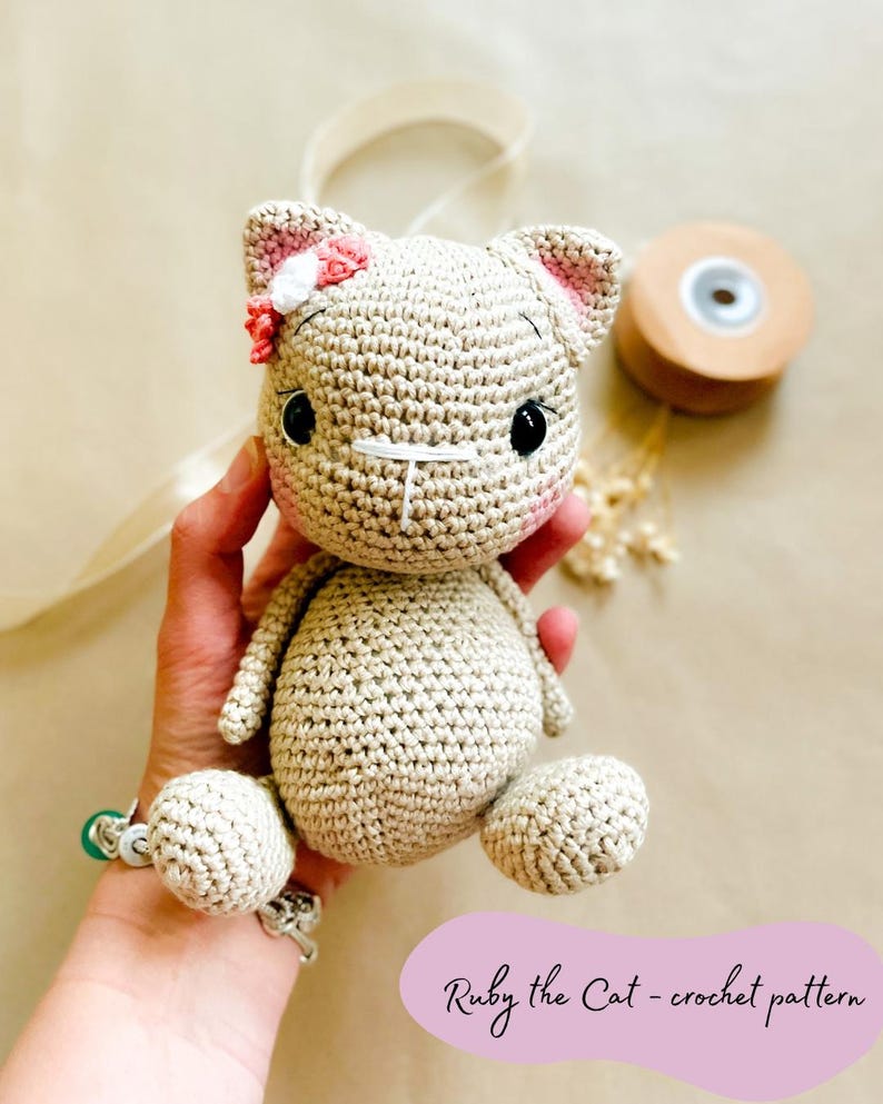 May include: A crocheted cat doll in beige yarn, with pink accents on the ears and a small flower. The cat has black eyes and a stitched nose and mouth. The text "Ruby the Cat - crochet pattern" is visible.