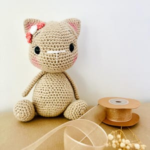May include: A crocheted cat doll in tan yarn with pink accents on the cheeks and ears. The cat has black button eyes and a small flower on its head. A spool of gold ribbon and a length of sheer ribbon are next to the doll.