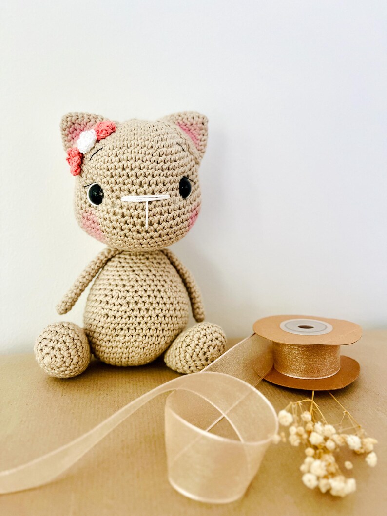 May include: A crocheted cat doll in tan yarn with pink accents on the cheeks and ears. The cat has black button eyes and a small flower on its head. A spool of gold ribbon and a length of sheer ribbon are next to the doll.