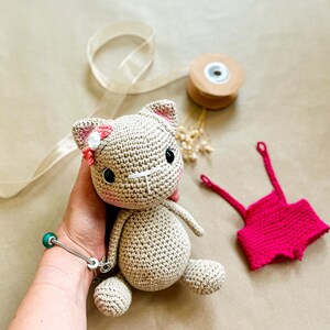 May include: A crocheted cat doll with a pink bow and flower on its head. The cat is beige with black eyes and a white nose. A small pair of pink crocheted shorts are next to the cat. The cat is approximately 6 inches tall.