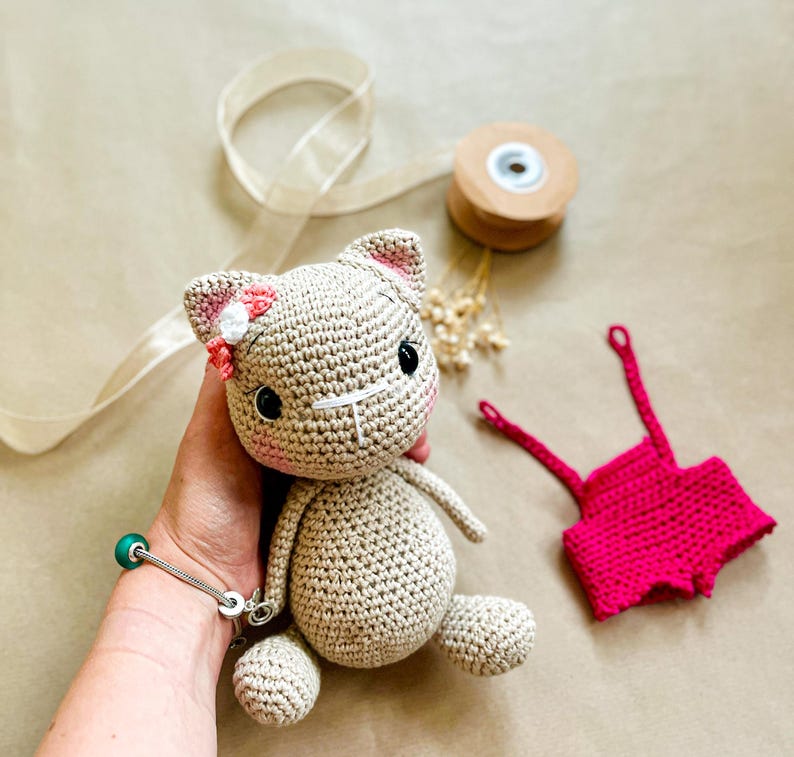 May include: A crocheted cat doll with a pink bow and flower on its head. The cat is beige with black eyes and a white nose. A small pair of pink crocheted shorts are next to the cat. The cat is approximately 6 inches tall.