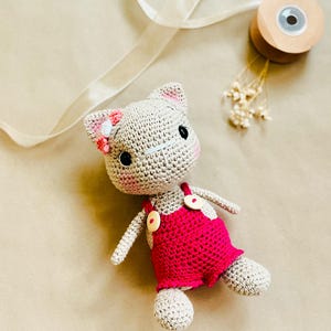 May include: A handmade crochet cat doll with a light beige body and pink accents. The cat wears a bright pink overall dress with wooden buttons. The cat has black eyes, pink cheeks, and a flower on its ear. The doll is lying on a neutral surface.