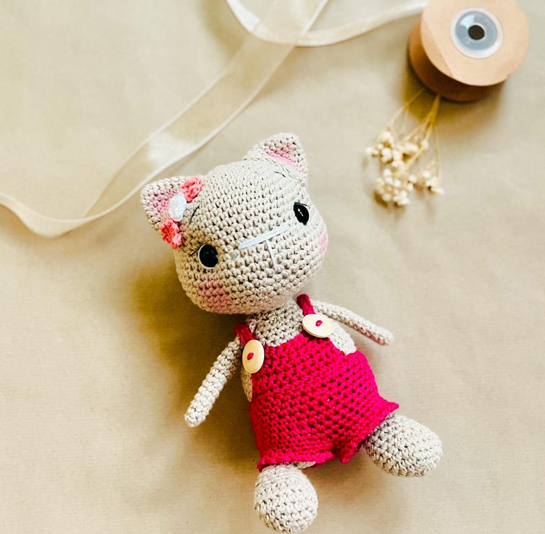 May include: A handmade crochet cat doll with a light beige body and pink accents. The cat wears a bright pink overall dress with wooden buttons. The cat has black eyes, pink cheeks, and a flower on its ear. The doll is lying on a neutral surface.