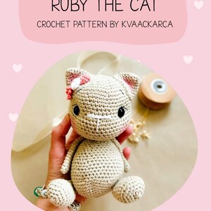May include: A crocheted cat doll with the text "RUBY THE CAT" and "CROCHET PATTERN BY KVAACKARCA". The cat is beige with pink accents on the ears and a pink bow. The doll has black eyes and is sitting. The background is pink.