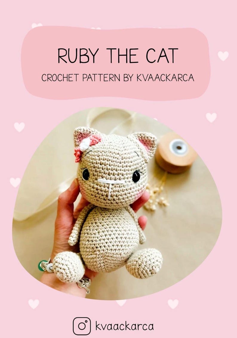 May include: A crocheted cat doll with the text "RUBY THE CAT" and "CROCHET PATTERN BY KVAACKARCA". The cat is beige with pink accents on the ears and a pink bow. The doll has black eyes and is sitting. The background is pink.