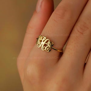 Può includere: Un anello dorato con un design a monogramma. L'anello è indossato su un dito.