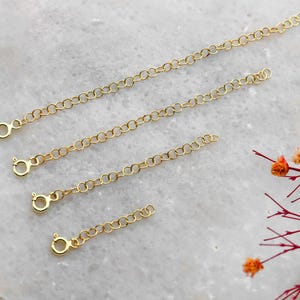 May include: Five gold chain extenders of varying lengths, laid out on a white marble surface. The extenders are made of delicate, linked chains with lobster claw clasps.
