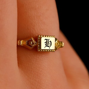 Puede incluir: Un anillo de sello de oro con un engaste cuadrado que presenta una letra "H" negra en fuente gótica.