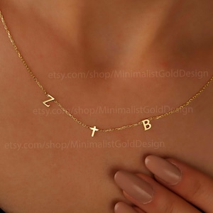 以下が含まれることがあります： Z、T、Bの文字が入った繊細なゴールドネックレス。ネックレスは人の首にかけられています。文字は小さく、間隔をあけて配置されています。背景は温かみのある肌の色です。ネックレスはミニマリストジュエリーです。