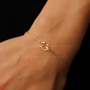 May include: A gold chain bracelet with a delicate charm featuring the number "13" in a stylized heart shape.