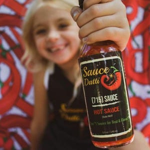 May include: A glass bottle of hot sauce with a black label that says "Sauce Datti (715) Sauce Hot Sauce Shake Well One Source for Heat & Flavor 5 fl oz / 148 ml". The bottle is being held by a person's hand.