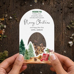 May include: An arched white card featuring watercolor illustrations of green pine trees, pinecones, and wrapped gifts. The text reads: WISHING YOU holiday cheer, prosperity, peace and abundant growth. Merry Christmas and Happy New Year. LOVE, ASHLEY.