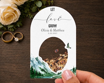 May include: An arched white card featuring the text LET love GROW, Olivia & Matthew, and 22.5.2027. The card has a circular cutout filled with seeds, and a watercolour illustration of green pine trees and blue mountains at the bottom.