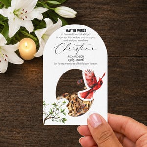 May include: A white arched memorial card featuring a watercolor red cardinal in flight, a tree branch, and text that reads: MAY THE WINDS of heaven blow and whisper in your ear that we love and miss you, and wish you were here. Christine RICHARDSON 1965-2026.
