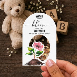 May include: An arched white card featuring the black text WATCH THESE bloom JUST LIKE BABY WREN WHO WILL BE HERE SOON! and a watercolor illustration of a pink flower growing from brown soil with green leaves, with a circular cutout in the center.