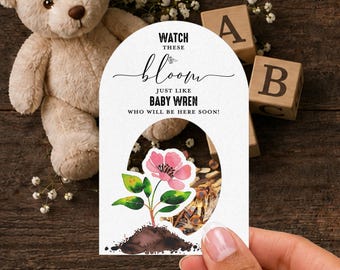 Puede incluir: Una tarjeta blanca arqueada con el texto negro WATCH THESE bloom JUST LIKE BABY WREN WHO WILL BE HERE SOON! y una ilustración en acuarela de una flor rosa creciendo desde tierra marrón con hojas verdes, con un recorte circular en el centro.