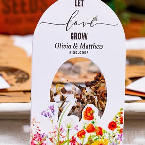 May include: A white arched paper seed packet featuring a clear window filled with seeds. The text reads LET love GROW, Olivia & Matthew, and 5.22.2027. The bottom is decorated with a colorful illustration of various wildflowers including red poppies.