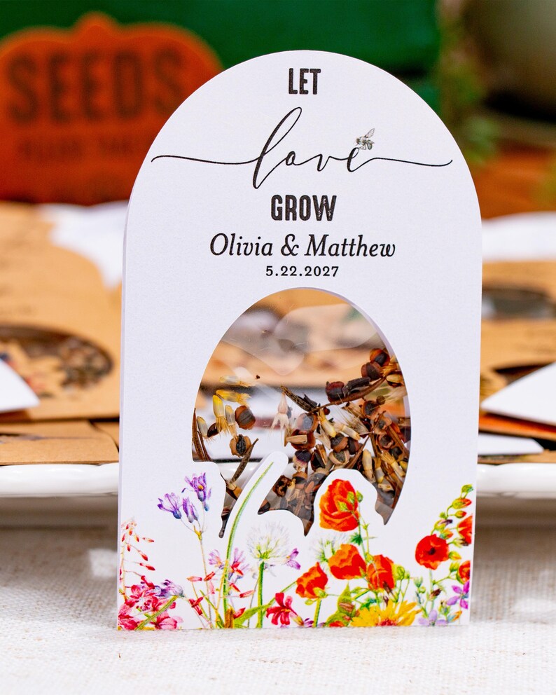 May include: A white arched paper seed packet featuring a clear window filled with seeds. The text reads LET love GROW, Olivia & Matthew, and 5.22.2027. The bottom is decorated with a colorful illustration of various wildflowers including red poppies.