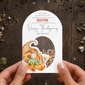 May include: An arched white card featuring a watercolor cornucopia filled with pumpkins, sunflowers, and autumn leaves. The text reads: Just like a garden blooms with nourishment and care, may our GRATITUDE for one another grow wildly each year. Happy Thanksgiving. The Millers.