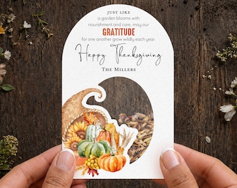 May include: An arched white card featuring a watercolor cornucopia filled with pumpkins, sunflowers, and autumn leaves. The text reads: Just like a garden blooms with nourishment and care, may our GRATITUDE for one another grow wildly each year. Happy Thanksgiving. The Millers.