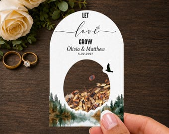 Könnte beinhalten: Eine bogenförmige weiße Papierkarte mit dem Text LET love GROW, Olivia & Matthew und 5.22.2027. Ein kreisförmiger Ausschnitt zeigt braune Samen im Inneren. Unten ist eine Aquarell-Wald- und Seenlandschaft mit einem kleinen schwarzen Vogel.
