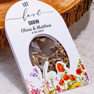May include: A white arched seed packet featuring the text LET love GROW, Olivia & Matthew, and 5.22.2027. The packet has a clear oval window displaying brown seeds and is decorated with colorful illustrations of red, yellow, and purple wildflowers.