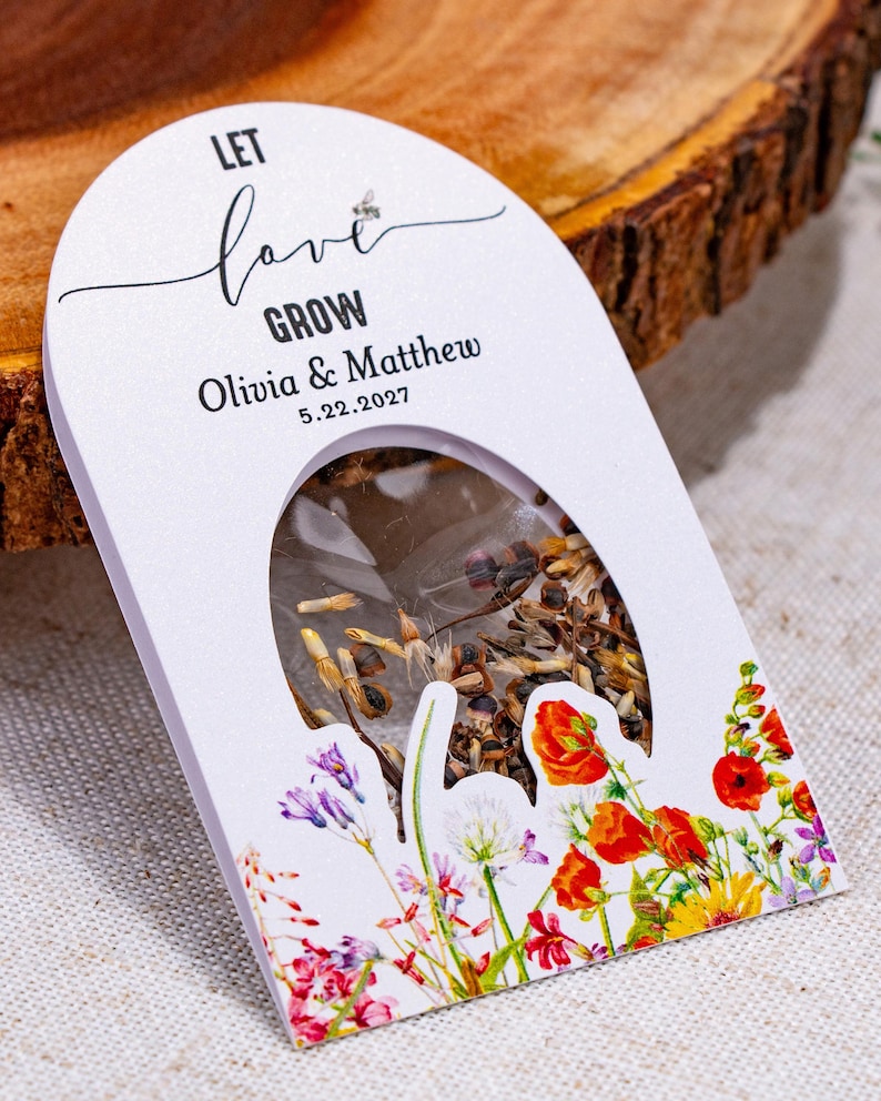 May include: A white arched seed packet featuring the text LET love GROW, Olivia & Matthew, and 5.22.2027. The packet has a clear oval window displaying brown seeds and is decorated with colorful illustrations of red, yellow, and purple wildflowers.