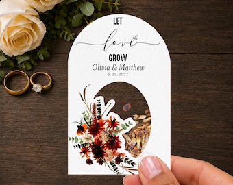 May include: An arched white card with the text LET love GROW, Olivia & Matthew, and 5.22.2027. The card features a floral illustration with red, orange, and brown flowers and green leaves, alongside a circular cutout revealing seeds inside.