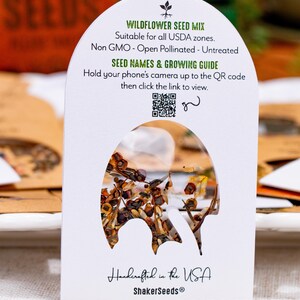 May include: A white arched seed packet labeled Wildflower Seed Mix with text stating Non GMO, Open Pollinated, Untreated, and a QR code for a growing guide. The packet features a central cutout window displaying a mix of brown and tan seeds.