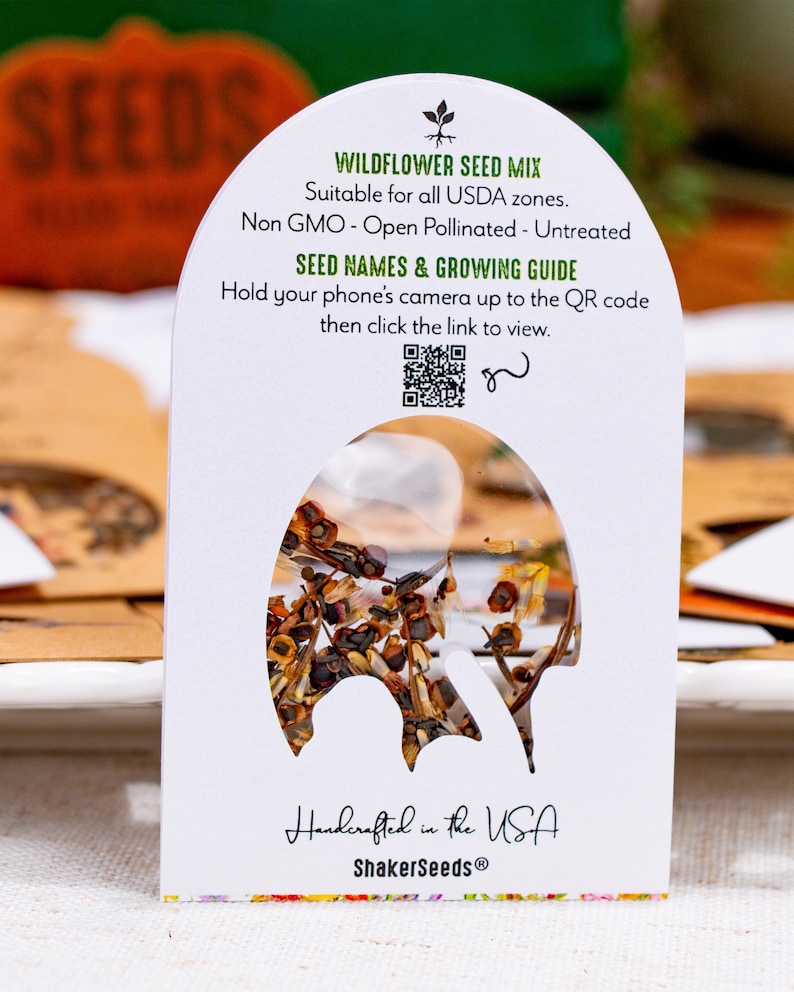 May include: A white arched seed packet labeled Wildflower Seed Mix with text stating Non GMO, Open Pollinated, Untreated, and a QR code for a growing guide. The packet features a central cutout window displaying a mix of brown and tan seeds.