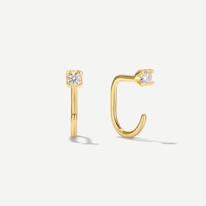 Puede incluir: Un par de pendientes de aro dorados con una pequeña piedra preciosa redonda y transparente engastada en cuatro garras en la parte superior de cada poste curvado. Los pendientes tienen un acabado pulido y un diseño abierto de líneas sencillas.