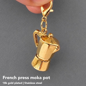 May include: A gold-plated stainless steel charm shaped like a moka pot coffee maker. The charm features a classic octagonal body, a handle, a lid, and a small knob on top. It is attached to a gold-coloured lobster claw clasp for easy attachment.