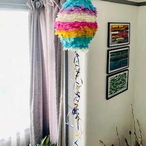 May include: A colourful rainbow pinata hanging from the ceiling with streamers. The pinata is made of tissue paper and is decorated with a variety of colours, including blue, pink, yellow, and green.