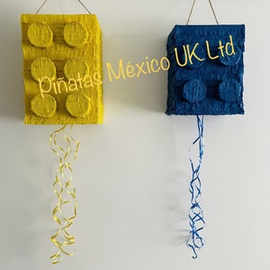 May include: Two Lego-shaped pinatas, one yellow and one blue, hanging from the ceiling with streamers. The yellow pinata has the text "Piñatas México UK Ltd" on it.