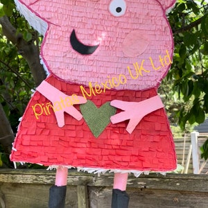 May include: A pink Peppa Pig pinata with a red dress and a green heart. The pinata has a yellow text label that says "Piñatas Mexico UK Ltd."