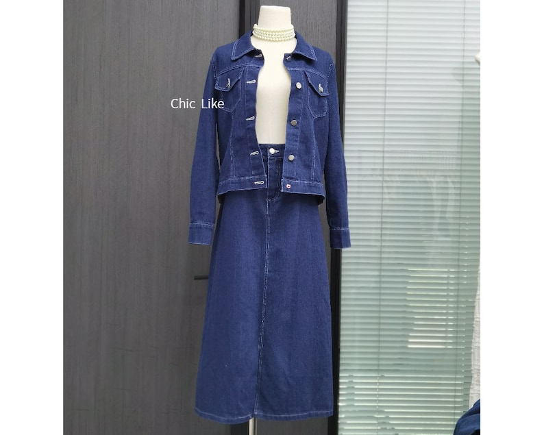 May include: A denim jacket and skirt set in a dark blue shade. The jacket has a classic collar, button closure, and two front pockets. The skirt is A-line and falls to the knee.