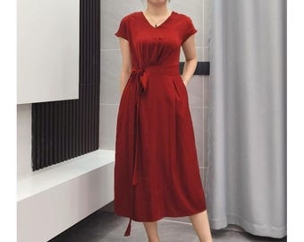 May include: A red midi dress with short sleeves and a V-neck. The dress features a tie-waist and pockets. The fabric appears to be a soft, flowing material. The dress is a solid color and suitable for various occasions.