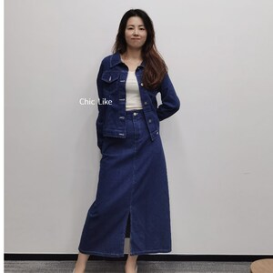 May include: A woman wearing a denim jacket and a denim skirt with a slit at the front. The jacket has a button fastening and the skirt has a high waist.