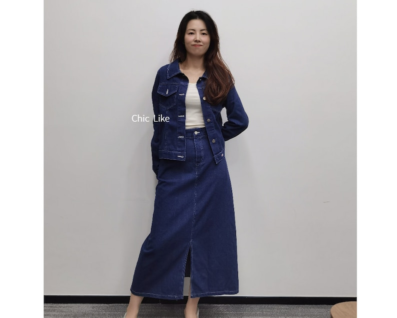 May include: A woman wearing a denim jacket and a denim skirt with a slit at the front. The jacket has a button fastening and the skirt has a high waist.