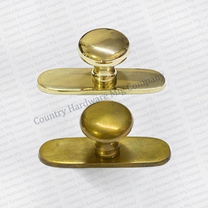 May include: Two brass door knobs with oval backplates. The knobs are round and have a smooth, polished finish. The backplates are rectangular and have a slightly textured surface.